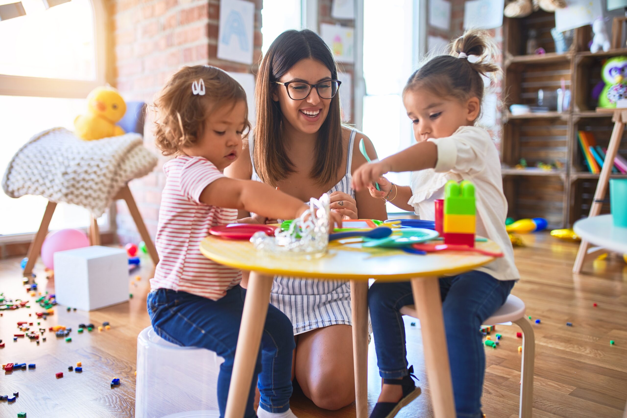 Young beautiful teacher and toddlers playing meals using plastic food and cutlery toy at kindergarten